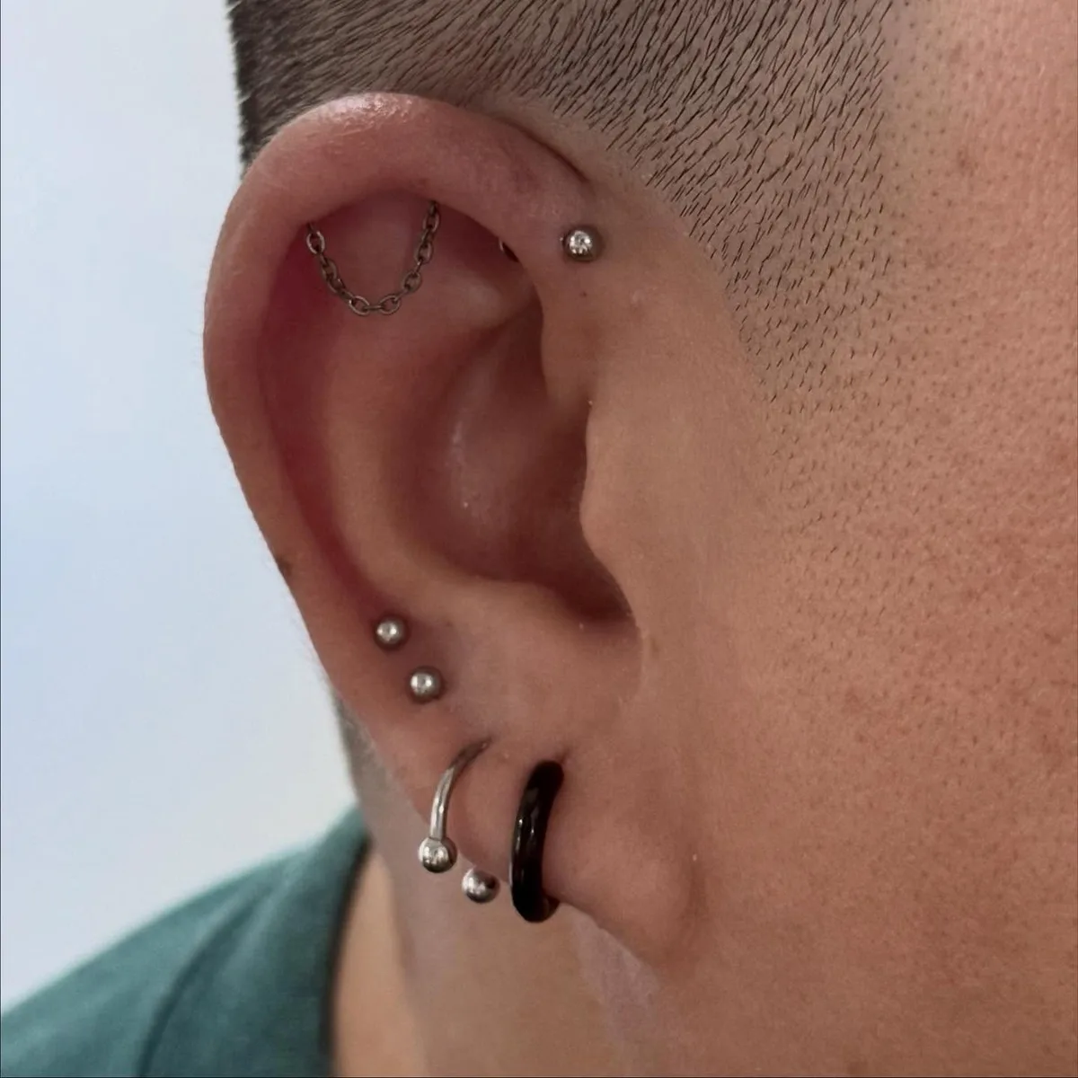 Silver chain linking two upper cartilage piercings, a forward helix stud, and stacked lobe studs and hoops