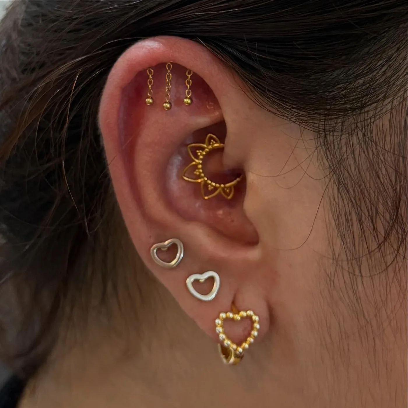 Curated ear with three gold chain-drop studs on the upper cartilage, a gold lotus daith ring, and heart studs on the lobe